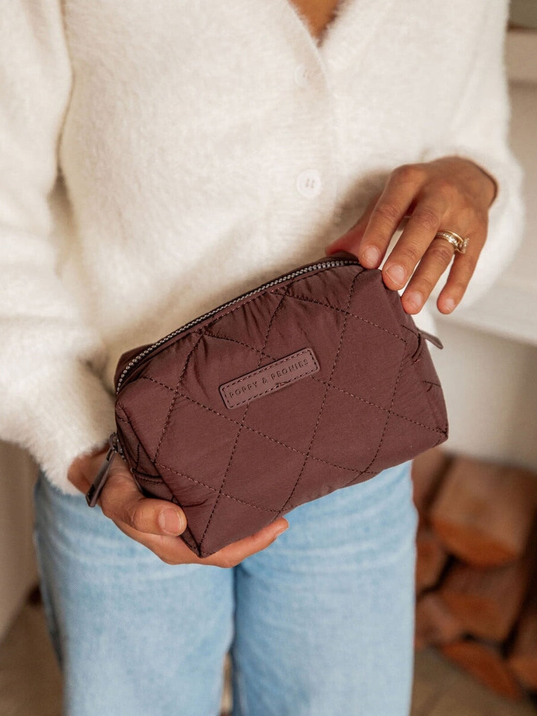 Person holding a quilted brown makeup bag with a brand logo, wearing a white sweater and light blue jeans.