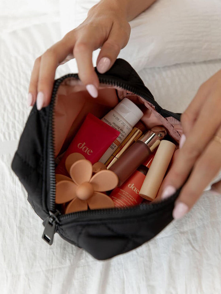 Black quilted makeup bag with various cosmetic products including lipsticks and a foundation stick on a white textured surface.