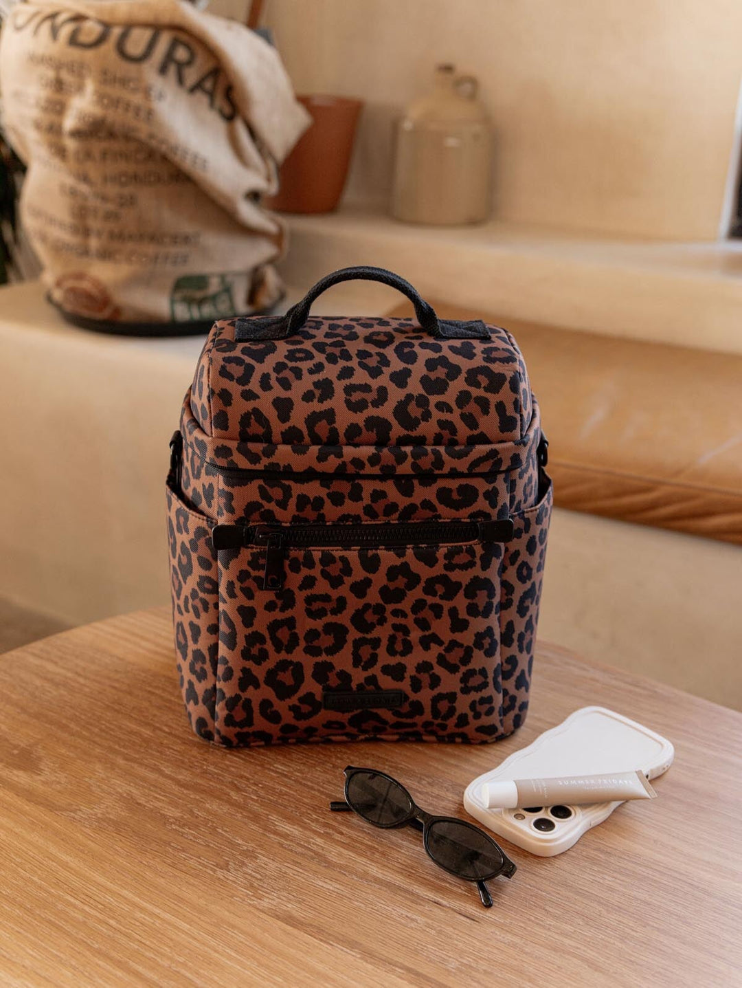 Leopard print lunch bag on a wooden surface with sunglasses and a phone case beside it.