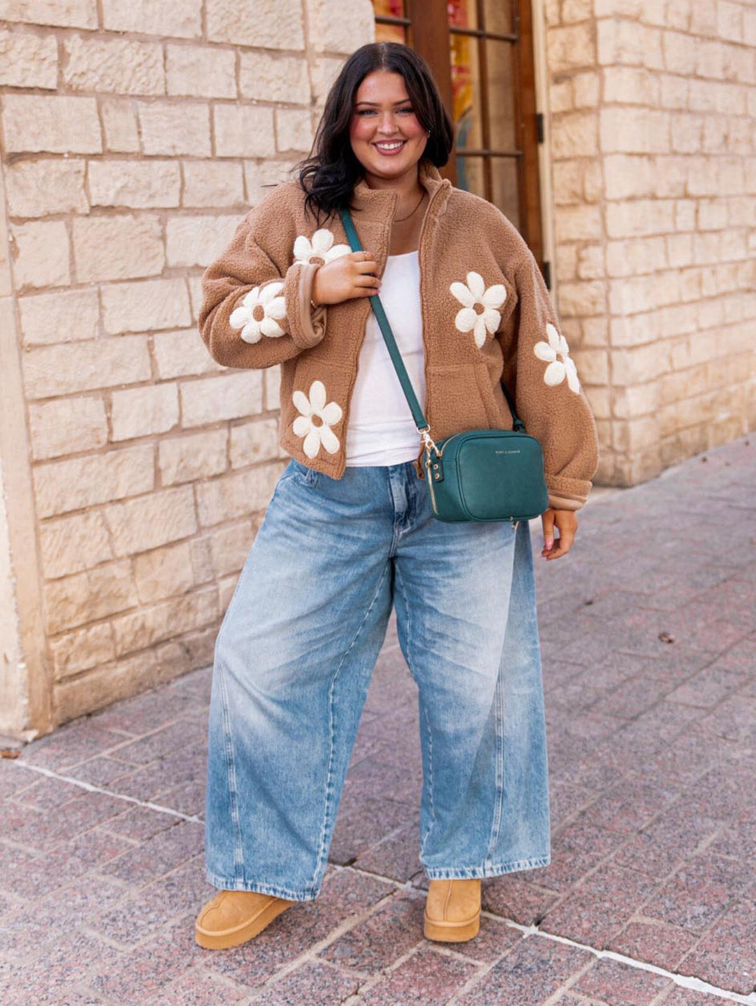Woman wearing a brown jacket with white floral patterns, white shirt, blue jeans, and tan shoes, holding a team crossbody bag.