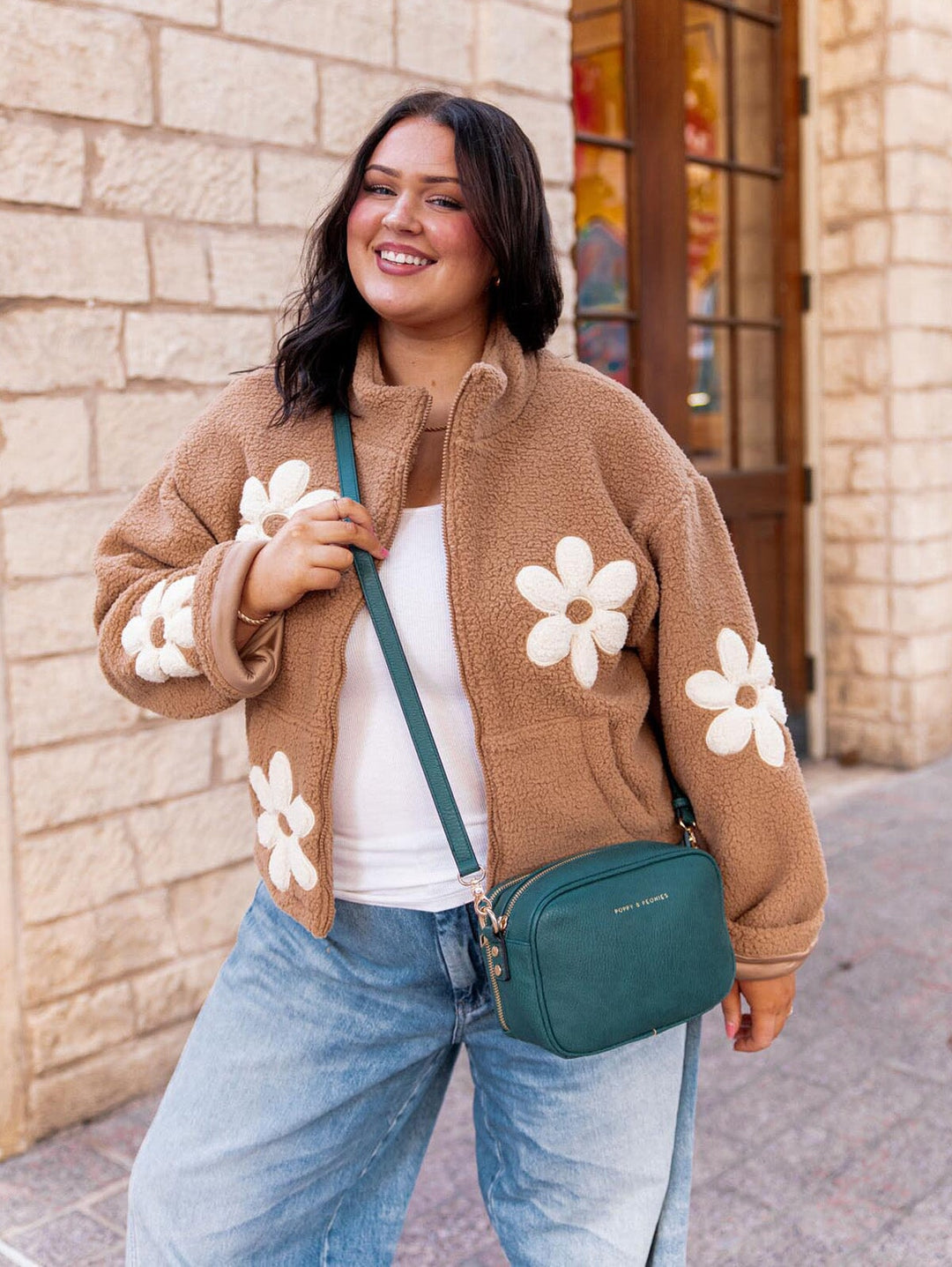 Woman wearing a brown jacket with white floral patterns, holding a teal crossbody bag, standing against a brick wall.