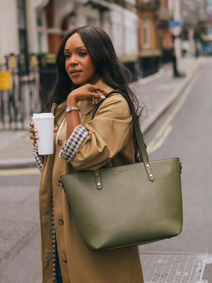 Woman wearing the Poppy & Peonies Go-Getter Tote in Olive over her shoulder while holding a takeaway coffee on a London street.