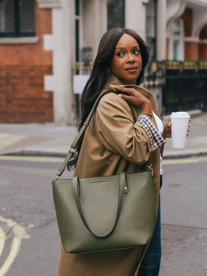 Woman carrying the Poppy & Peonies Go-Getter Tote in Olive over her shoulder while holding a takeaway coffee on a London street.