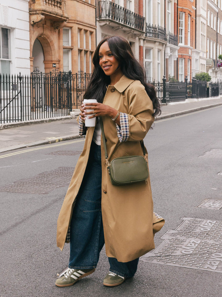 Full-body lifestyle shot of a woman wearing the Poppy & Peonies 24/7 Crossbody in Olive while walking along a city street with a takeaway coffee.