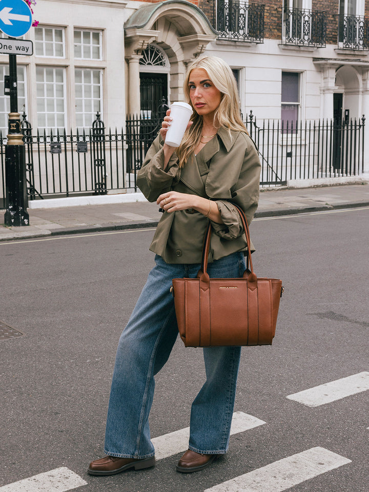 Woman carrying the Poppy & Peonies Small Multitasker in Chestnut by the top handles while holding a takeaway cup.