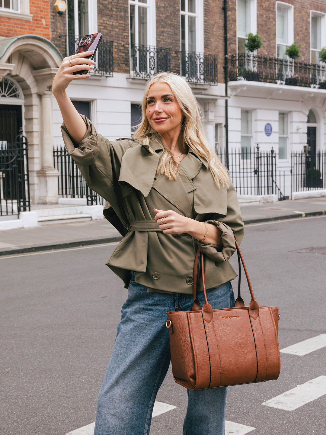 Woman carrying the Poppy & Peonies Small Multitasker in Chestnut by the top handles while taking a selfie on a city street.