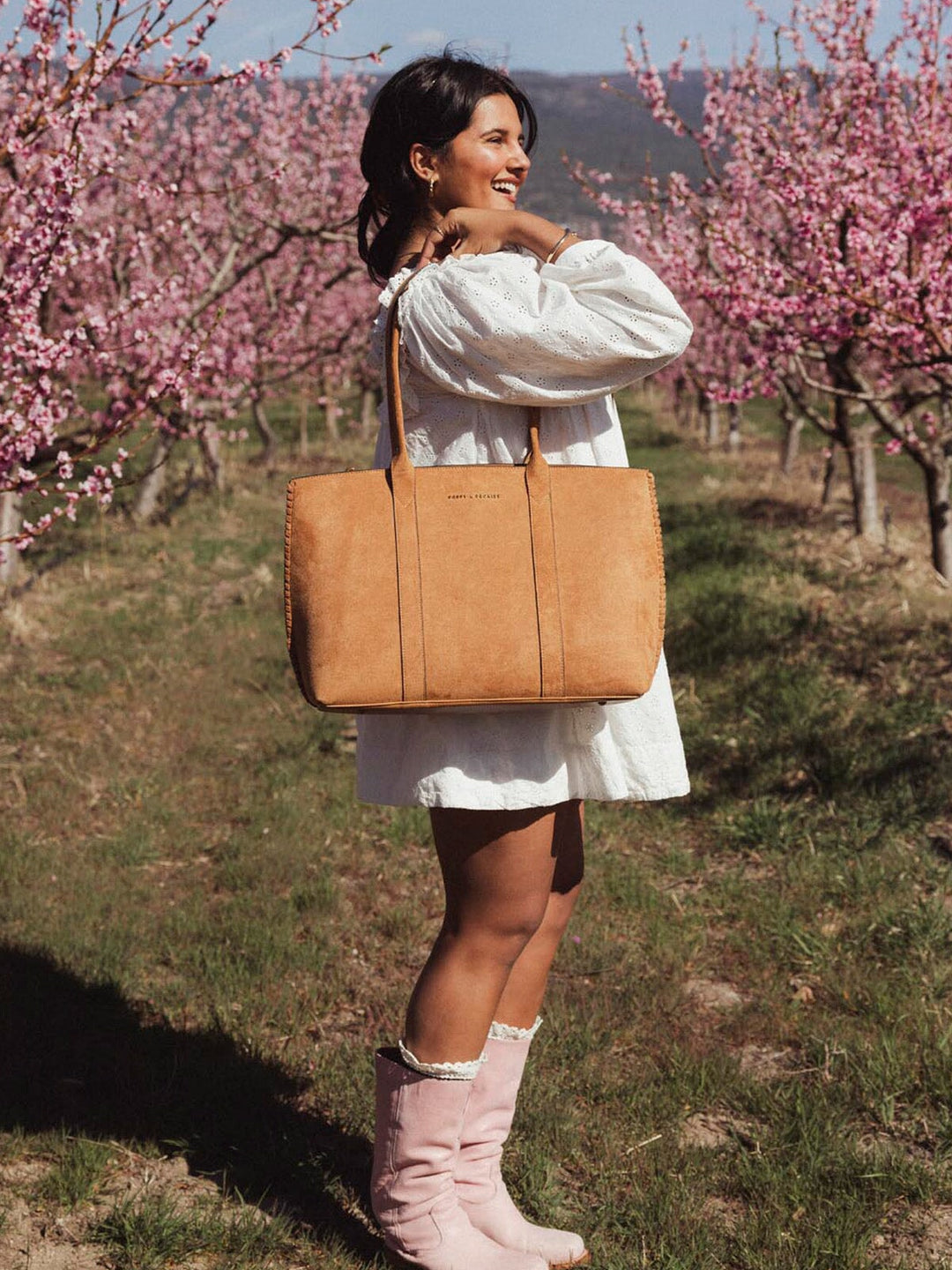 Woman in a white dress holding a brown vegan suede bag amongst cherry blossoms