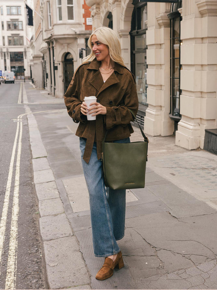 Full-body lifestyle shot of a woman wearing the Poppy & Peonies Bucket Bag in Olive over the shoulder while holding a takeaway coffee.
