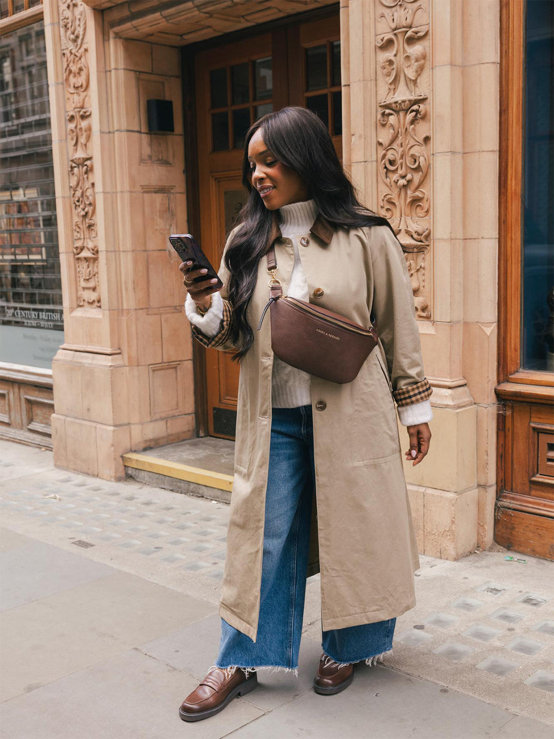 Full-body lifestyle shot of a woman wearing the Poppy & Peonies Sidekick in Espresso crossbody while checking her phone on a city street.