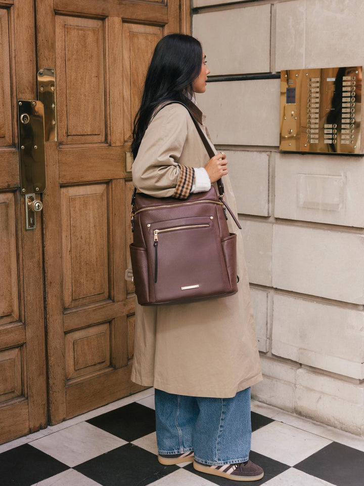 Woman wearing the Poppy & Peonies Commuter Backpack in Espresso over one shoulder while standing outside a city building.