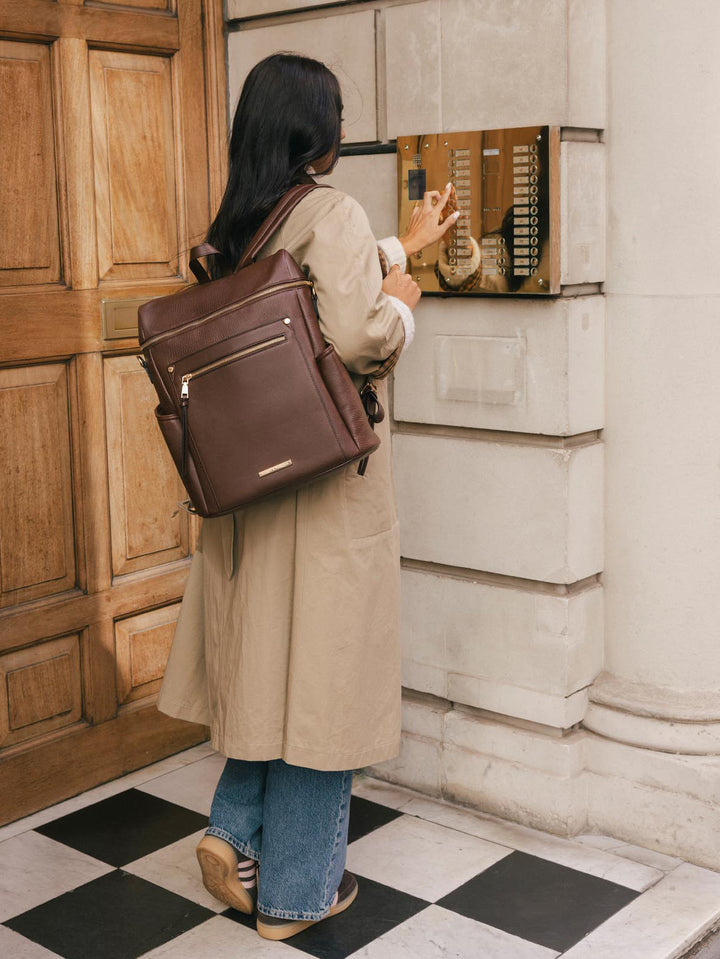 Woman wearing the Poppy & Peonies Commuter Backpack in Espresso over both shoulders while standing outside a city building.
