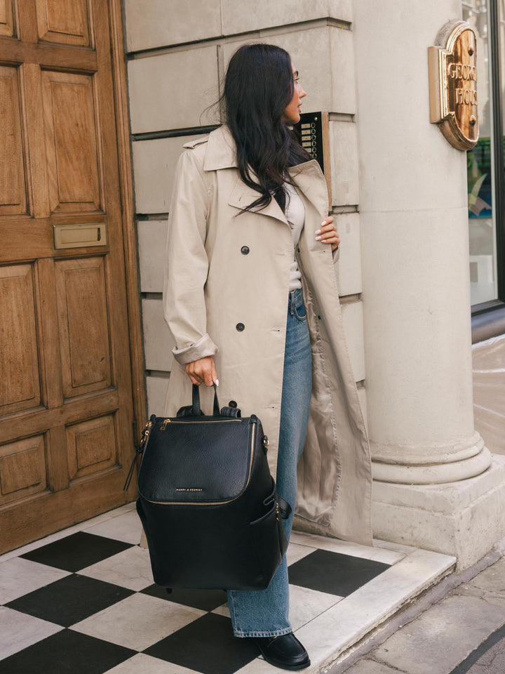Woman carrying the Poppy & Peonies Carryall Backpack in Black by the top handle while standing outside a city building.