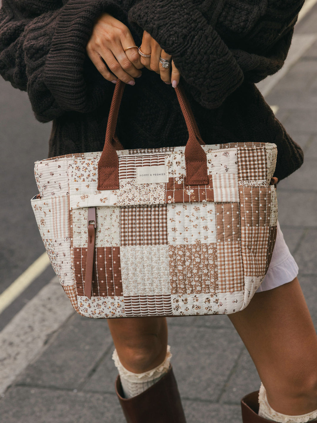 Close-up of a woman holding the Poppy & Peonies New Lunch Bag in Brown Patchwork by the top handles outdoors.