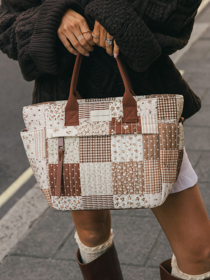 Close-up of a woman holding the Poppy & Peonies New Lunch Bag in Brown Patchwork by the top handles outdoors.