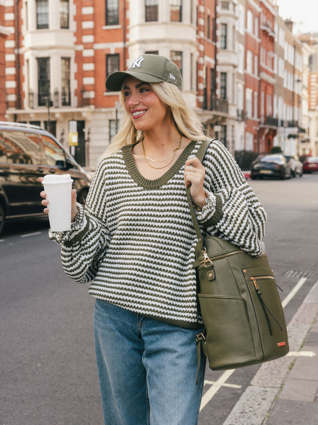 Woman carrying the Poppy & Peonies Commuter Backpack in Olive over one shoulder while smiling and holding a takeaway coffee.