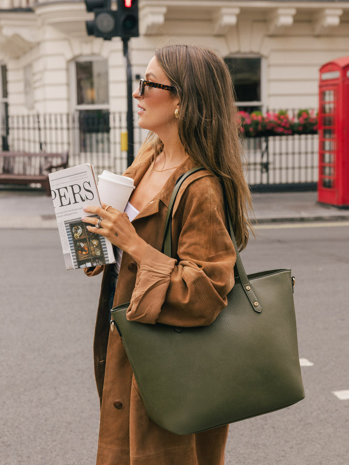 Woman carrying the Poppy & Peonies Go-Getter Tote in Olive over her shoulder while holding a takeaway coffee and magazine outdoors.