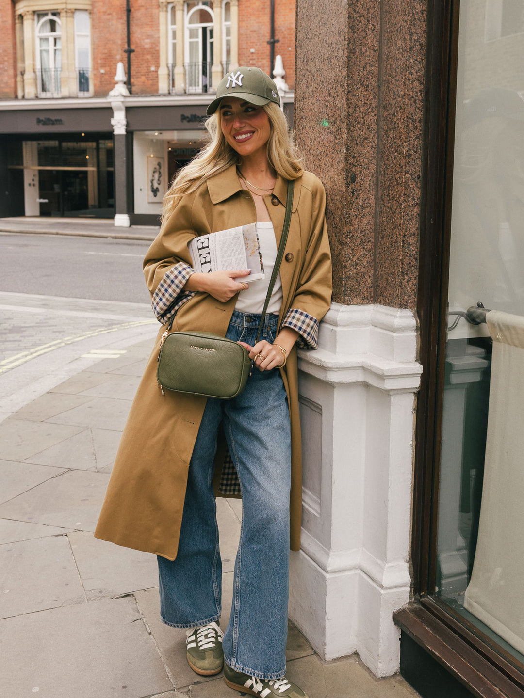 Full-body lifestyle shot of a woman wearing the Poppy & Peonies 24/7 Crossbody in Olive with a trench coat and jeans.
