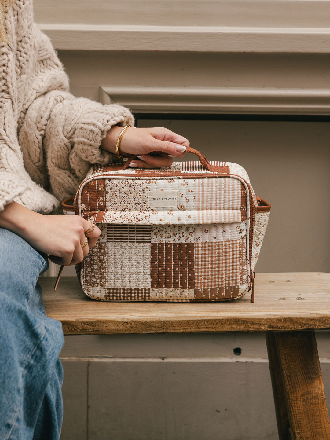 Close-up product-focused shot of the Poppy & Peonies Basic Lunch Bag in Brown Patchwork displayed on a bench, highlighting the patchwork print, top handle, and front pocket.