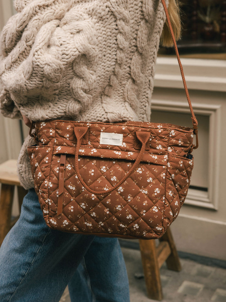 Close-up product-focused shot of the Basic Lunch Bag in Brown Floral worn over the shoulder, highlighting the quilted texture, handles, and front branding.