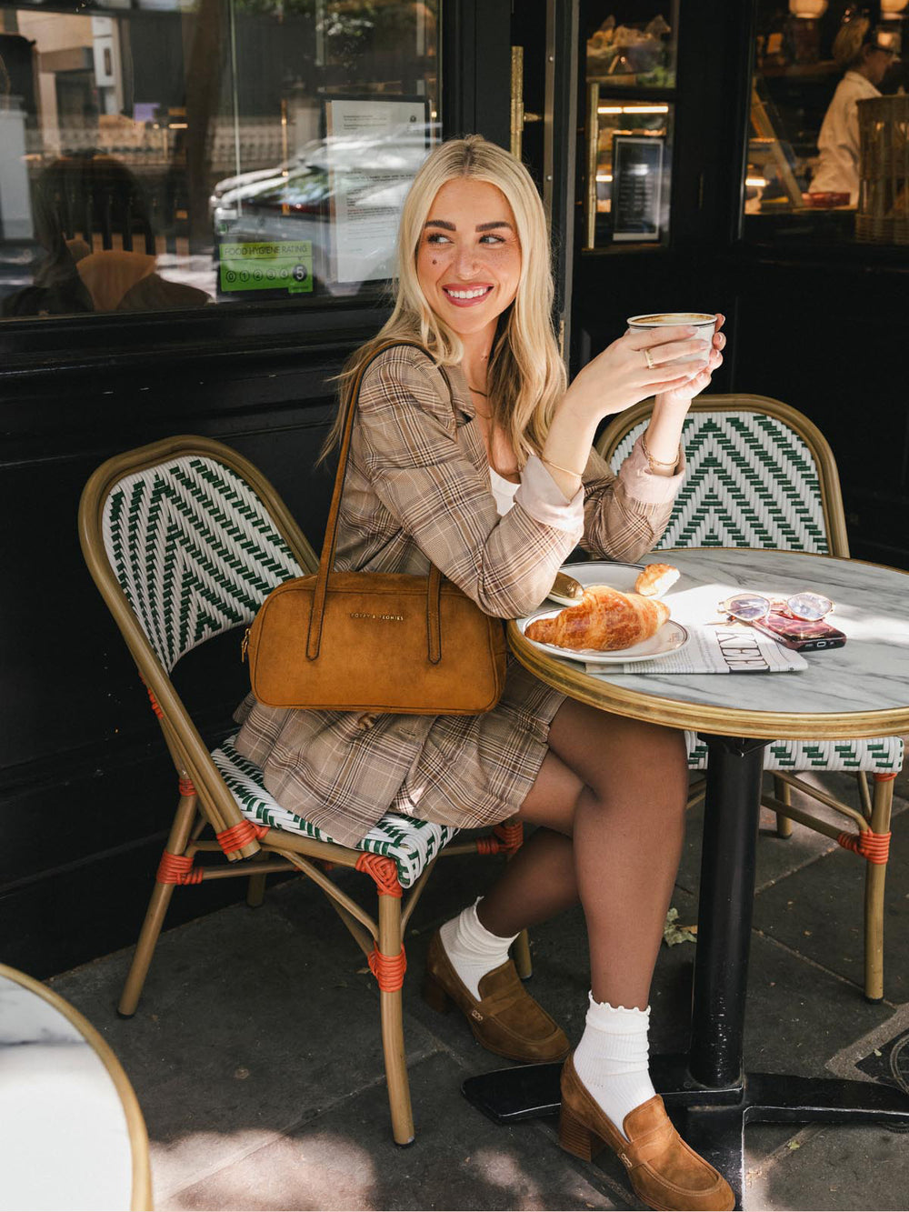 Poppy & Peonies The Friday Satchel in Cognac Suede carried by the top handles while seated at an outdoor café, highlighting its east-west silhouette and warm suede finish.