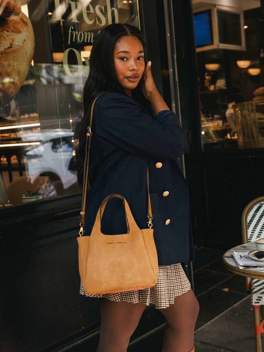 Poppy & Peonies The Pick Me Up Tote in Cognac Suede carried at the side outside a café, showcasing its top handles and everyday size.