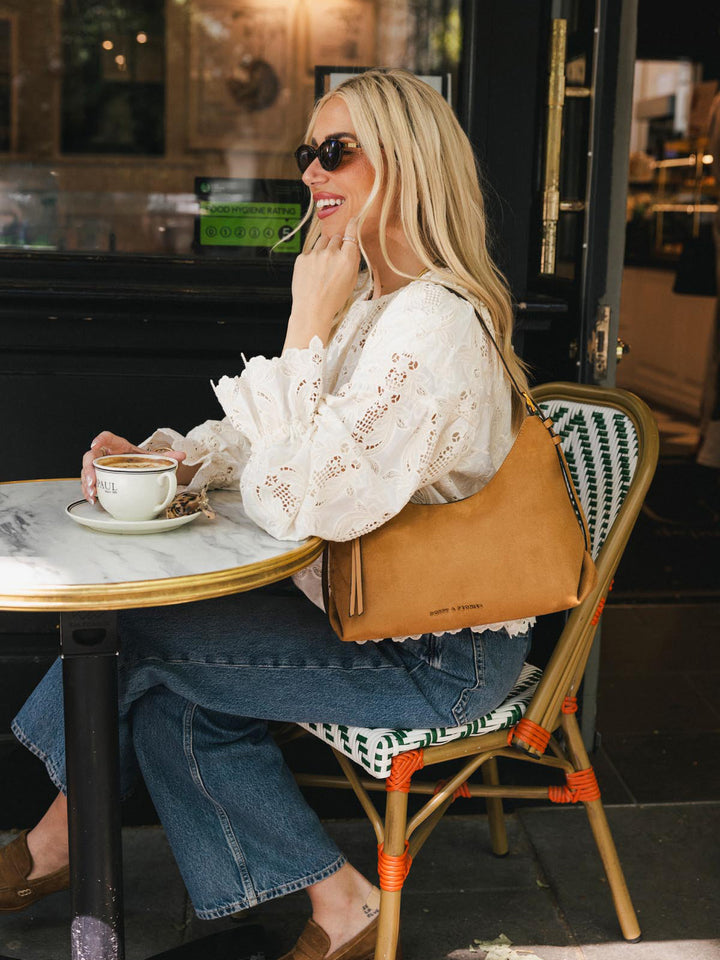 Poppy & Peonies The Sunday Bag in Cognac Suede worn over the shoulder while seated at an outdoor café.