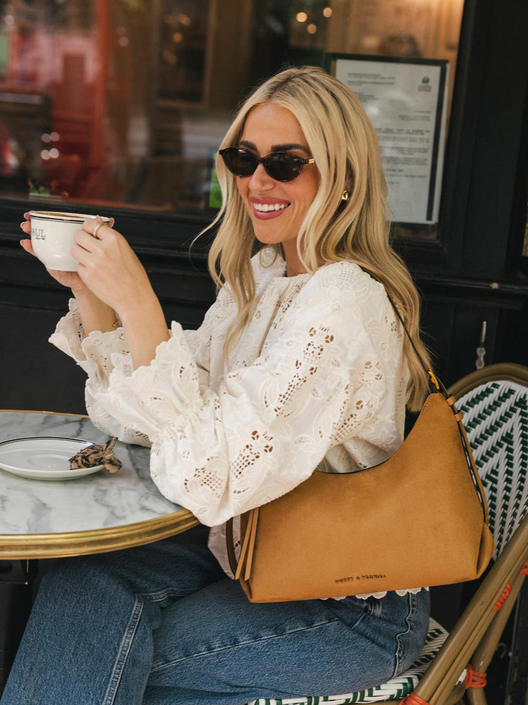 Poppy & Peonies The Sunday Bag in Cognac Suede worn over the shoulder at an outdoor café, highlighting its relaxed silhouette, suede finish and shoulder strap.
