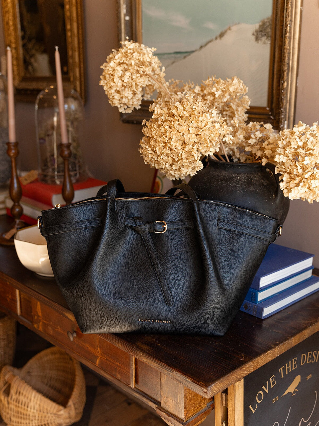 Poppy & Peonies The Knot Tote in Black displayed on a wooden table beside dried flowers, highlighting its spacious silhouette and long shoulder straps.