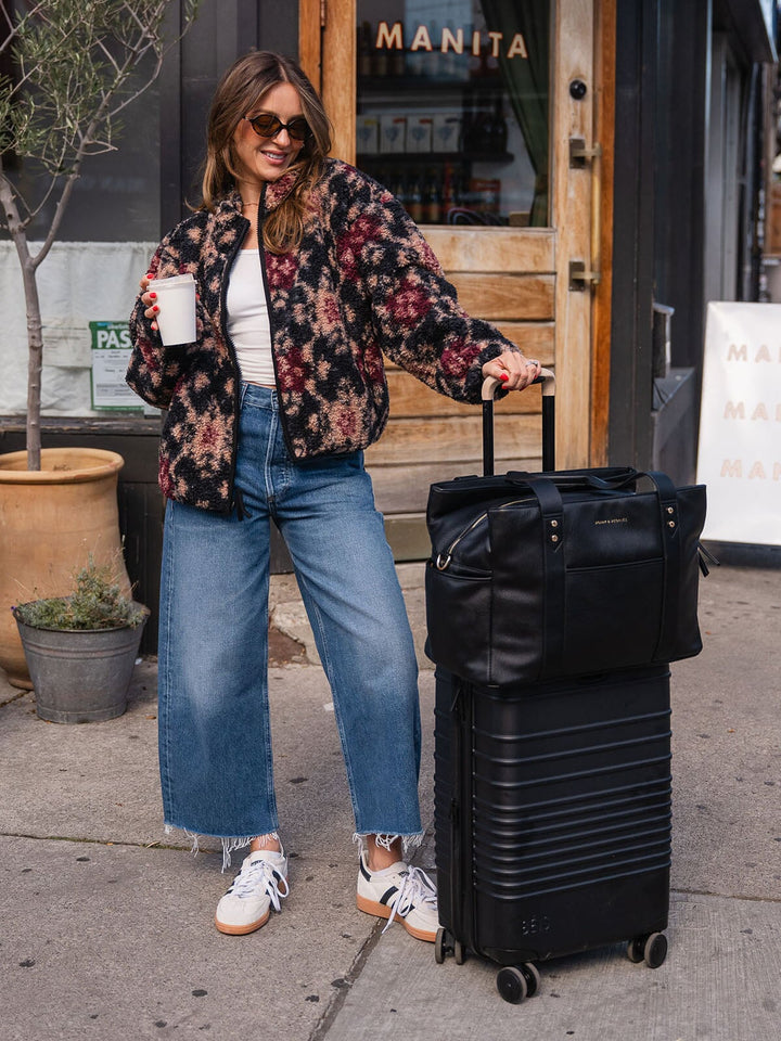 Woman standing with the Poppy & Peonies Mini Voyager in Black placed over the handle of a rolling suitcase. 
