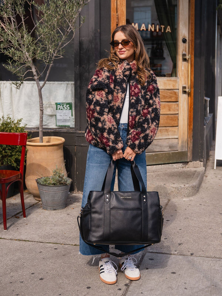 Woman carrying the Poppy & Peonies Mini Voyager in Black by the top handles while standing outdoors.