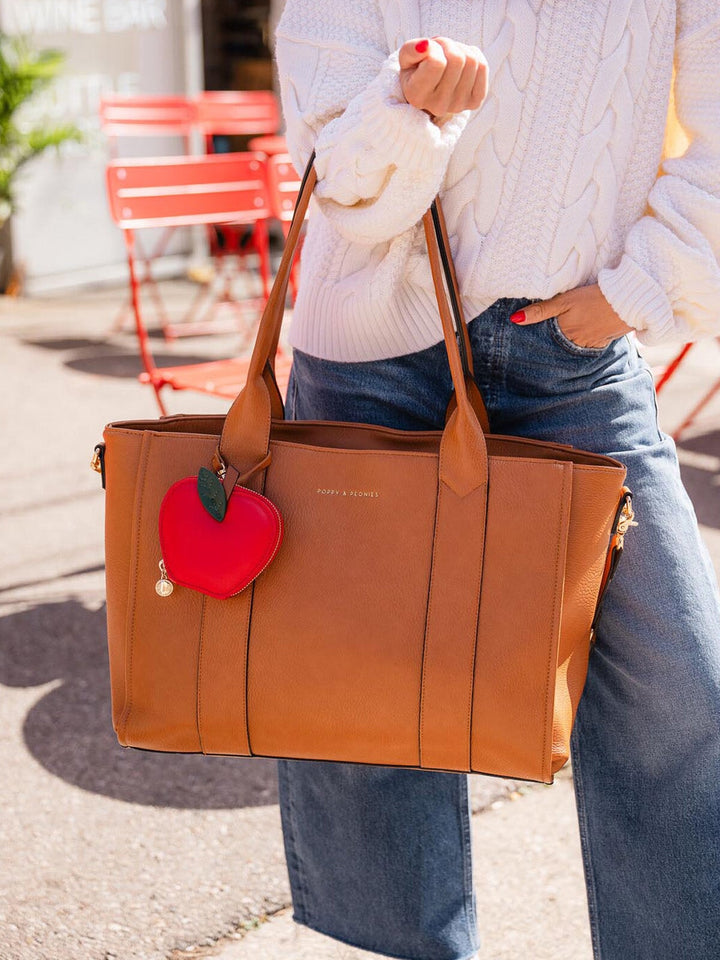 Person holding a brown tote bag with a red apple charm, wearing a white sweater and blue jeans.
