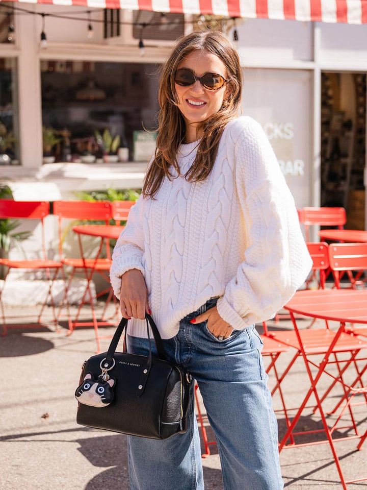 Woman in a white sweater and sunglasses holding a black handbag with a cat bag charm attached, standing in an outdoor setting with red tables and chairs.