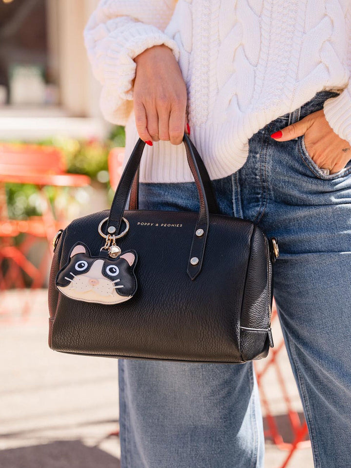 Person holding a black handbag with a cat charm, wearing a white sweater and blue jeans.