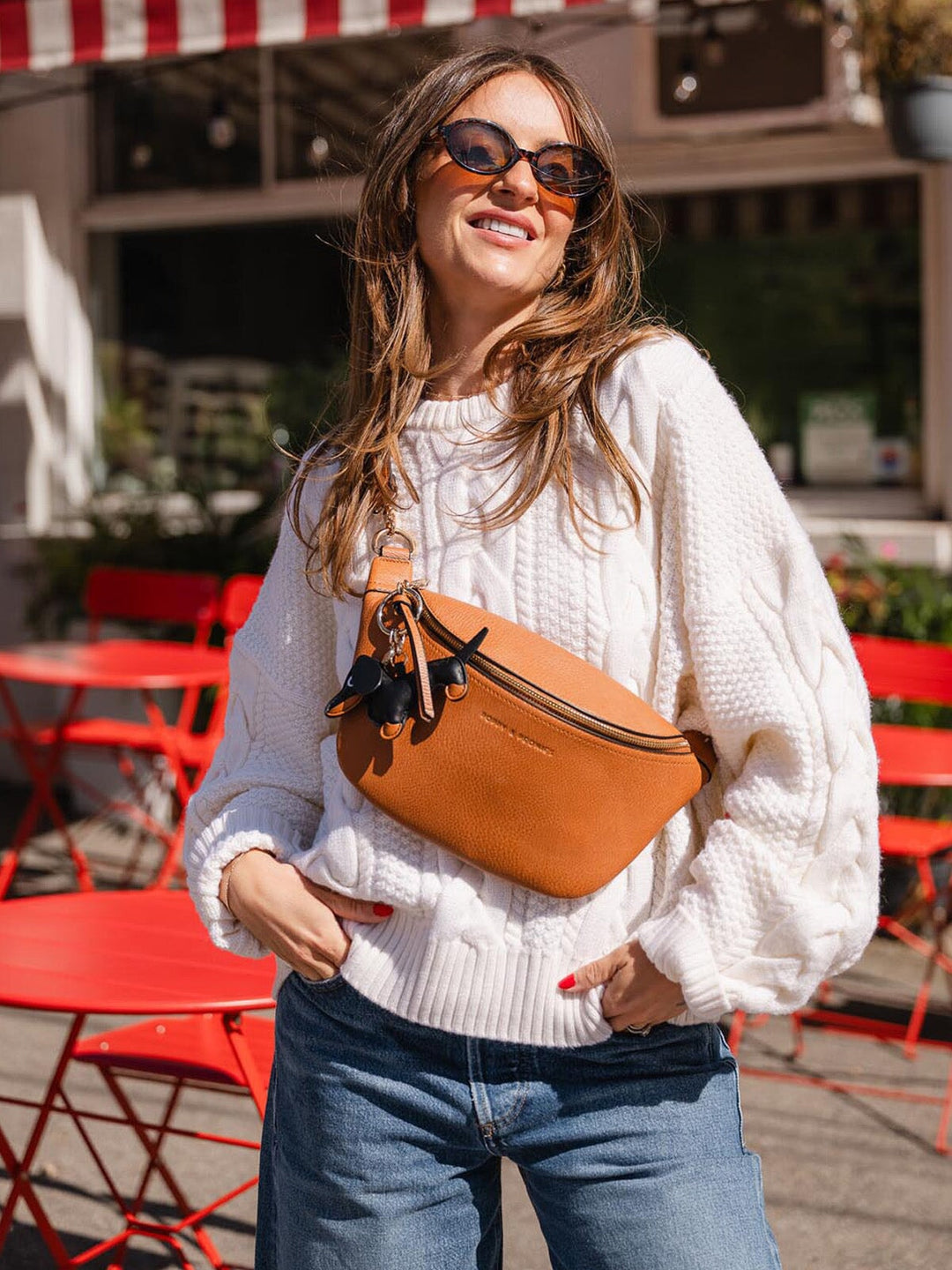 Woman wearing a white sweater and blue jeans, holding a brown bag with a dog bag charm attached.