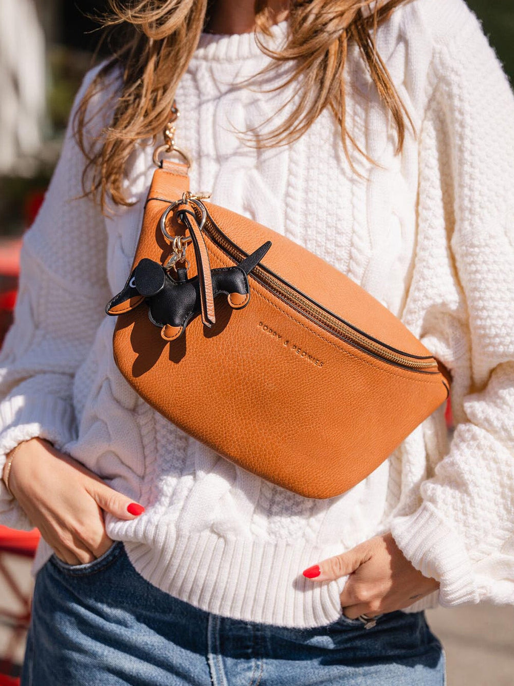 Person wearing a white sweater and blue jeans with a brown vegan leather bag with a Daschund bag charm.