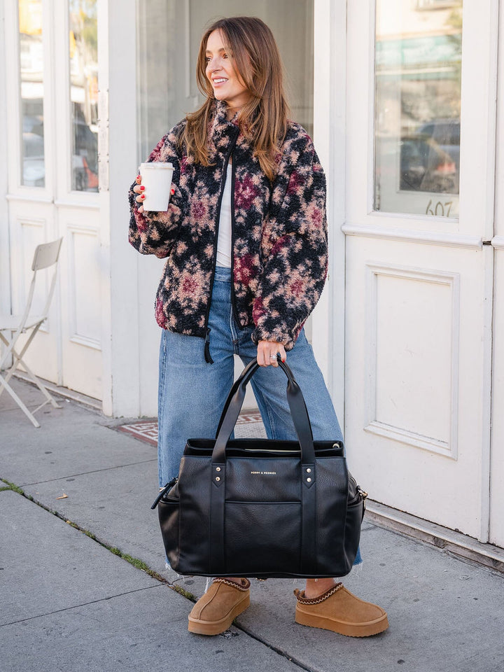 Woman carrying the Poppy & Peonies Mini Voyager in Black by the top handles while walking outdoors.