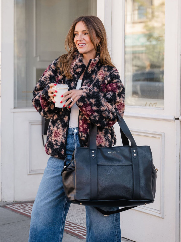 Woman carrying the Poppy & Peonies Mini Voyager in Black by the top handles while standing outdoors.