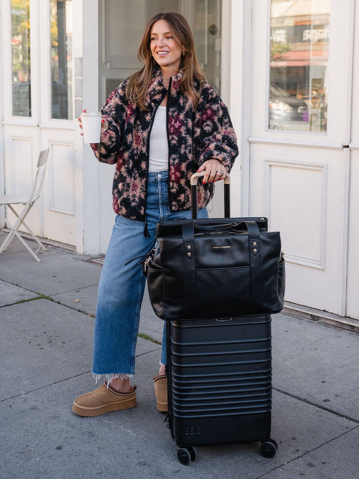 Woman posing with the Poppy & Peonies Mini Voyager in Black secured over the handle of a rolling suitcase.