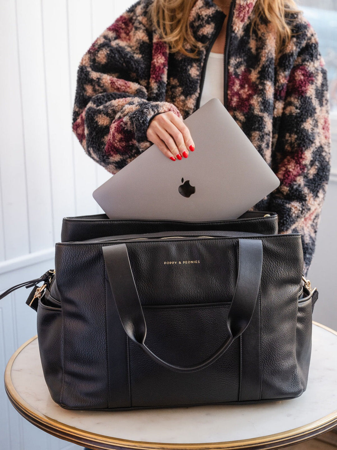 Lifestyle shot of the Poppy & Peonies Mini Voyager in Black, shown with a laptop being placed inside to highlight its spacious interior, laptop-friendly storage, its black exterior, front pocket, and sturdy shoulder straps.