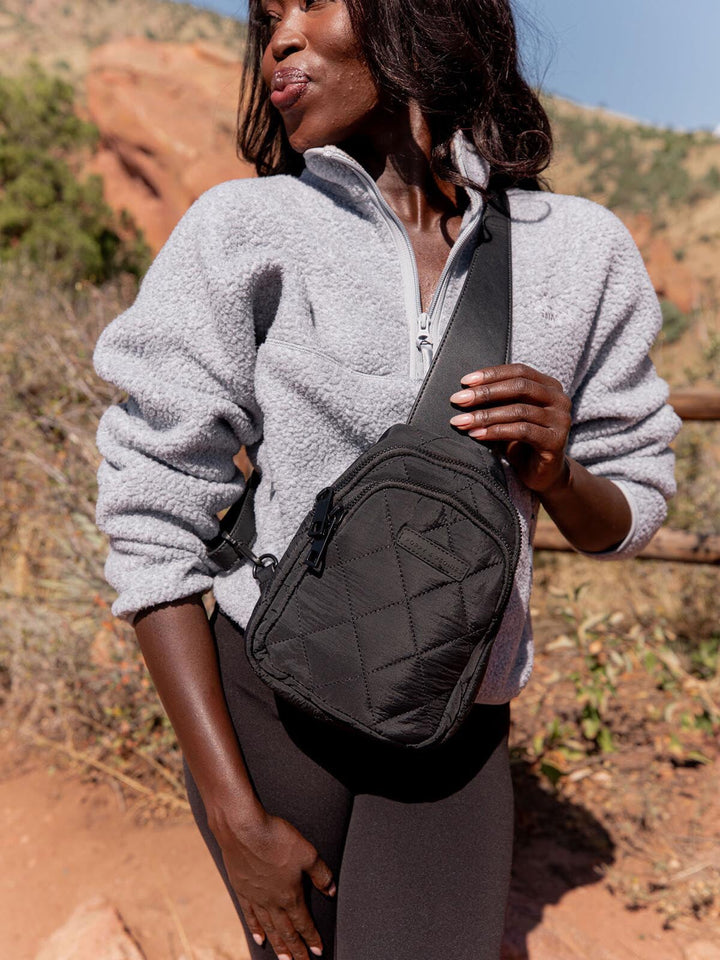 Person wearing a grey fleece jacket and holding a quilted black sling bag in a natural setting.