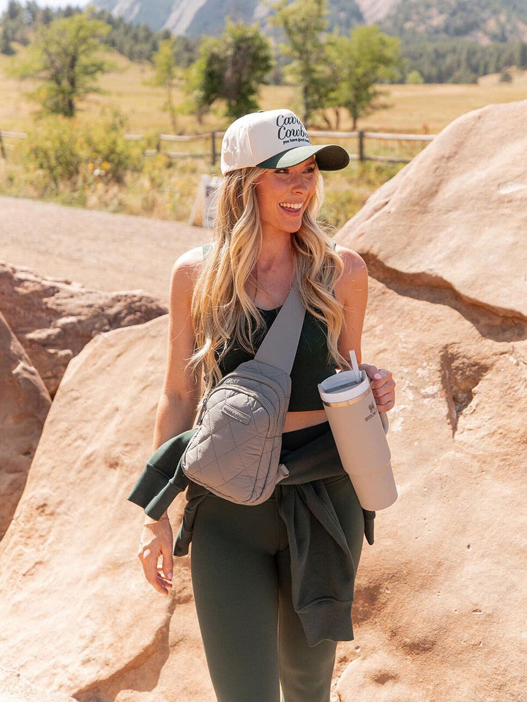 Woman in outdoor setting with mountains and trees, wearing a cap and carrying a grey quilted bag and tumbler.