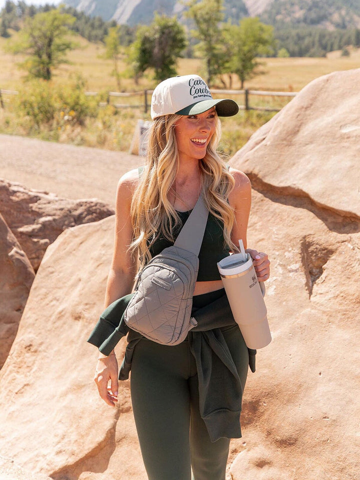 Woman in outdoor setting with mountains and trees, wearing a cap and carrying a grey quilted bag and tumbler.