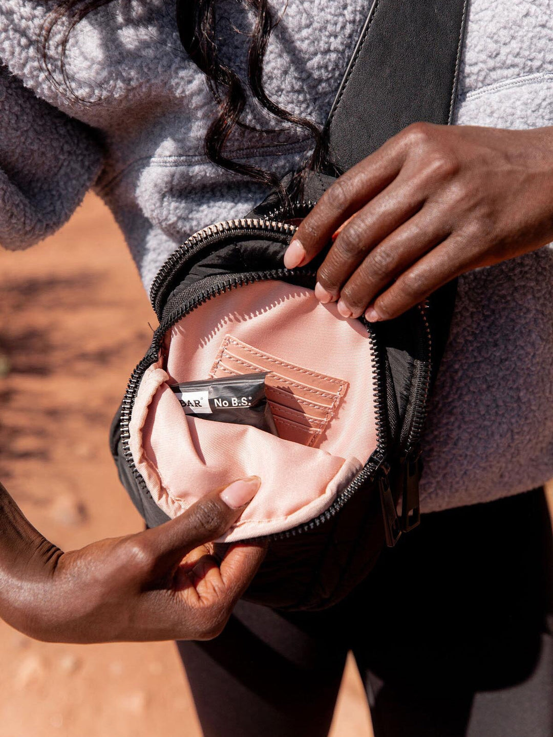 Person opening a black quilted crossbody bag with a pink interior, containing a small black object.