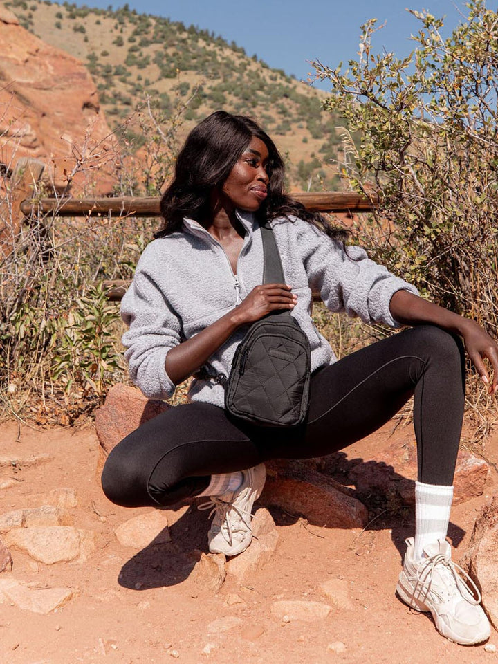 Woman wearing a black quilted sling bag sitting outdoors with a scenic background