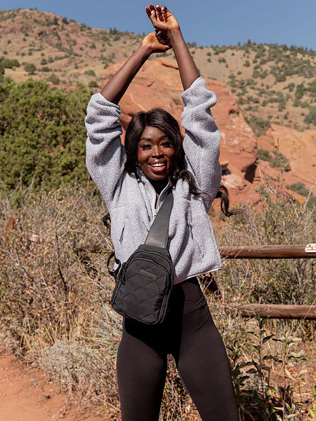 Person in grey jacket and black quilted sling bag with arms raised in a natural setting