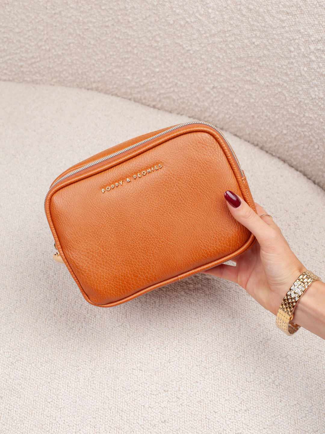 Handheld lifestyle shot of the Poppy & Peonies Pretty Connected Case in Cognac, showcasing its rich espresso exterior, compact structured silhouette, double-zip design, and gold-tone branding.