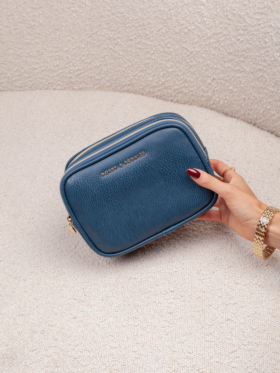 Handheld lifestyle shot of the Poppy & Peonies Pretty Connected Case in Navy, showcasing its rich navy exterior, compact structured silhouette, double-zip design, and gold-tone branding.