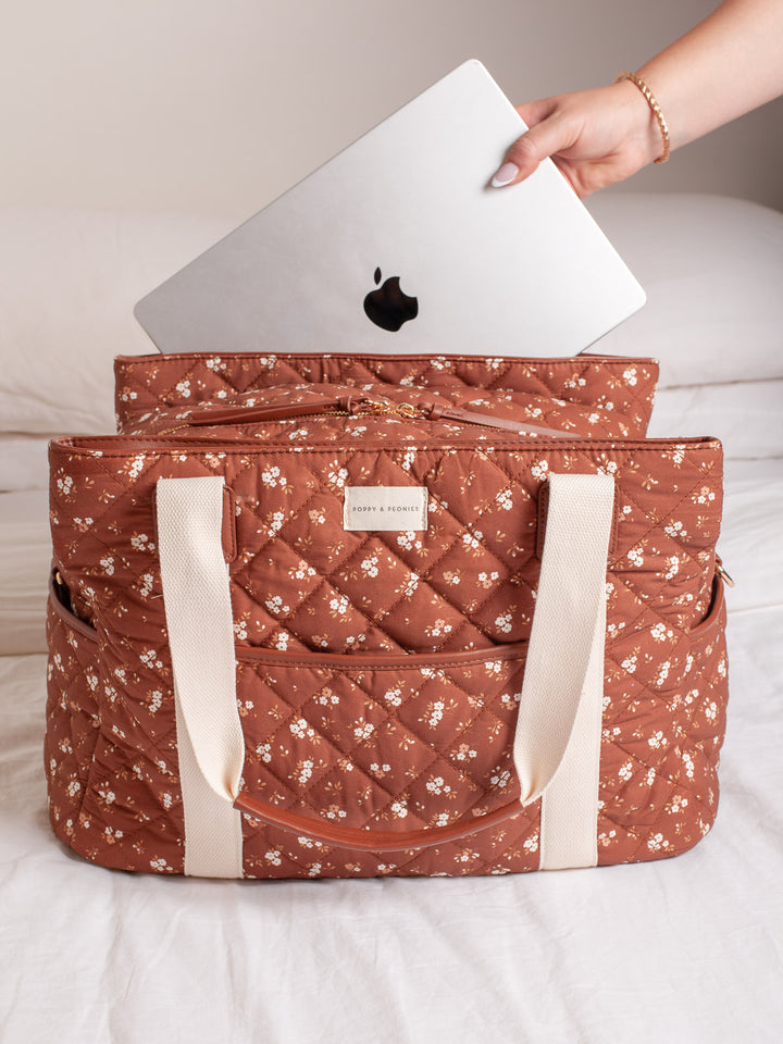 Lifestyle shot of the Poppy & Peonies Mini Voyager in Brown Floral, shown with a laptop being placed inside to highlight its spacious interior, laptop-friendly storage, quilted patchwork exterior, front pocket, and sturdy shoulder straps.