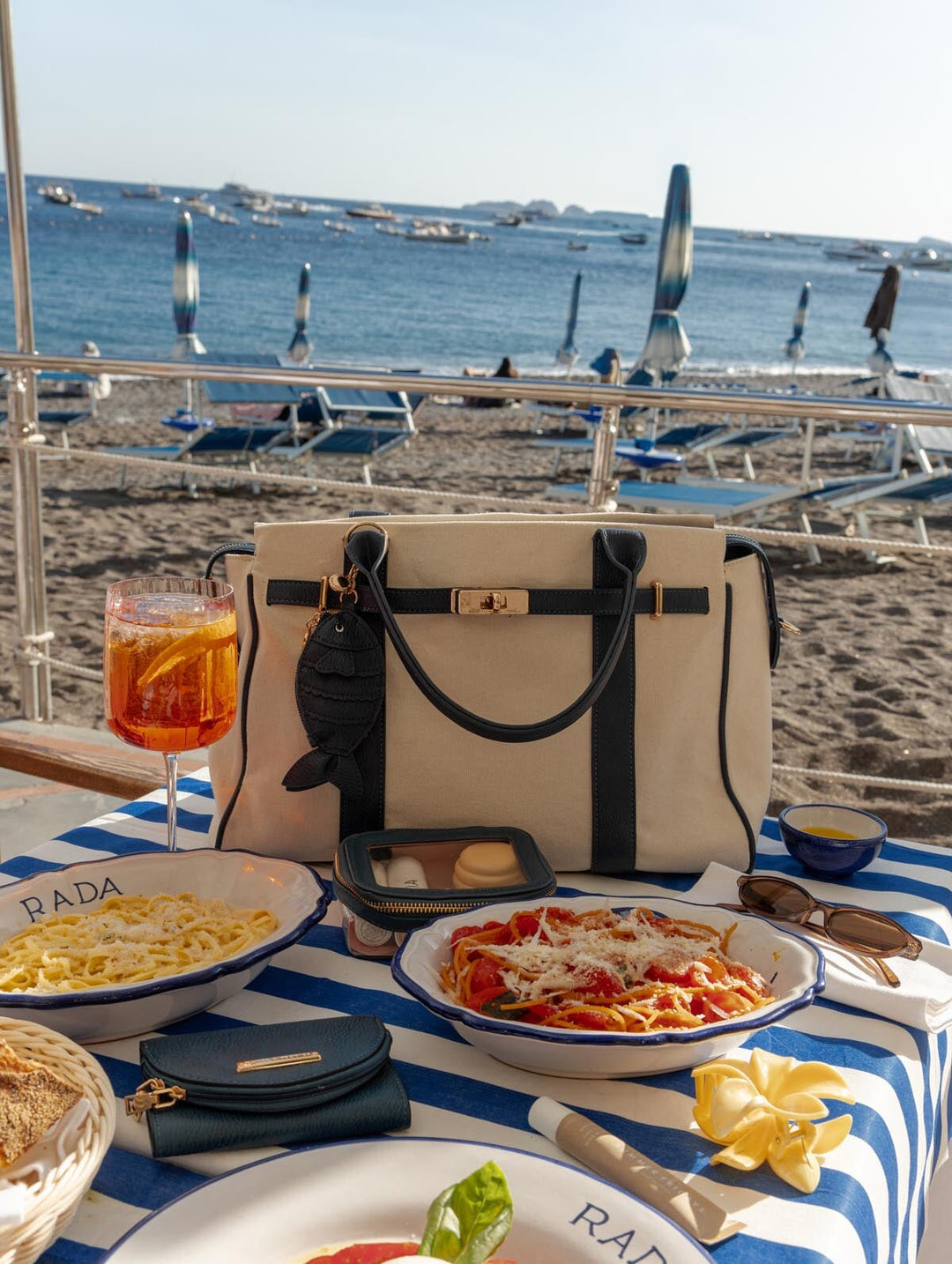 Canvas handbag with navy accents on a table by the beach with pasta dishes and drinks.
