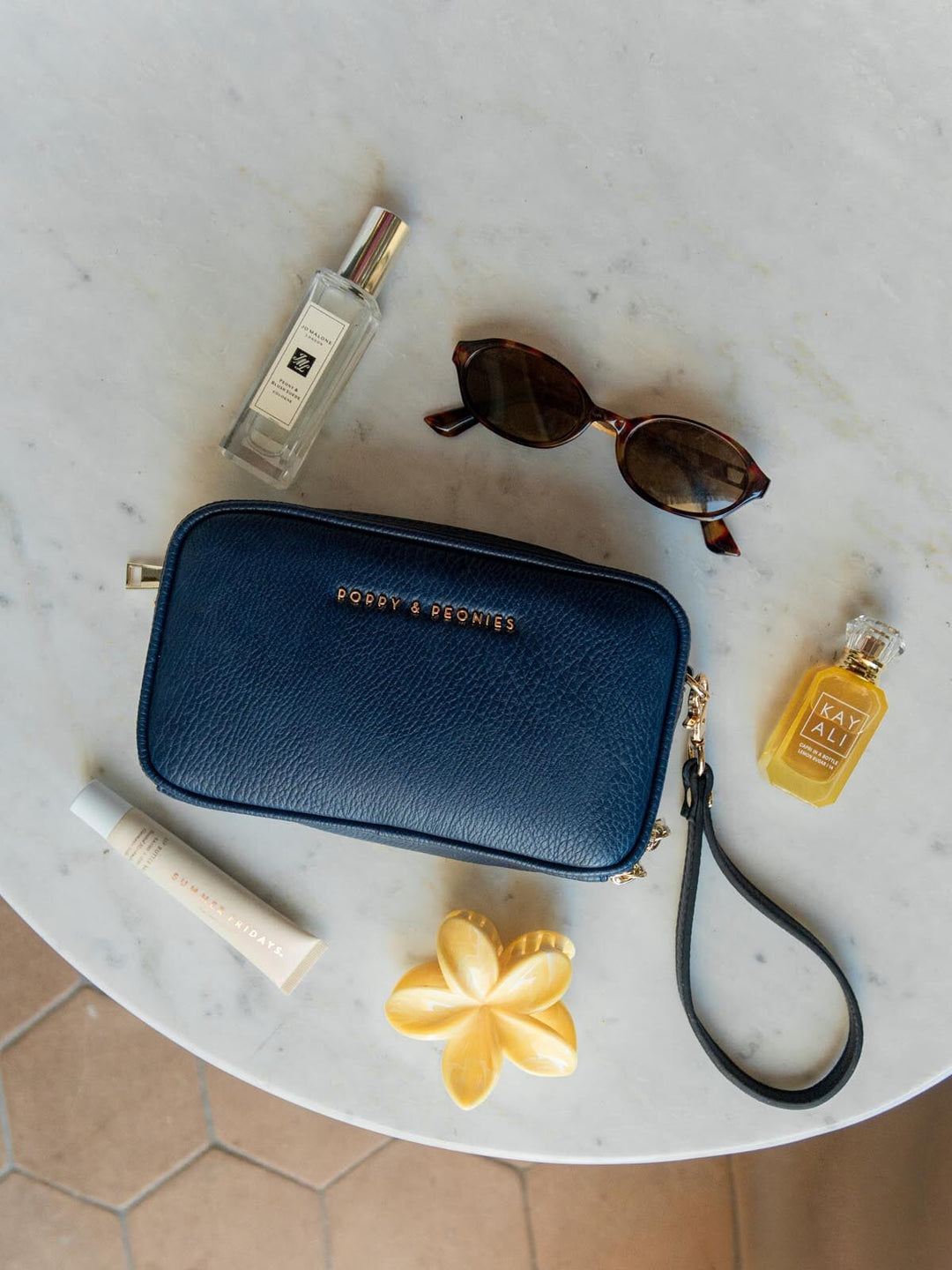 Blue handbag with Jo Malone perfume, sunglasses, and a yellow bottle on a marble surface.